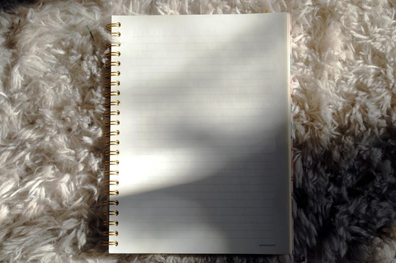 A small stack of contemporary essay collections beside a half-empty cup of coffee on a bright wooden table, with a paperback by Annie Ernaux on top, sunlight falling diagonally across the page edges.