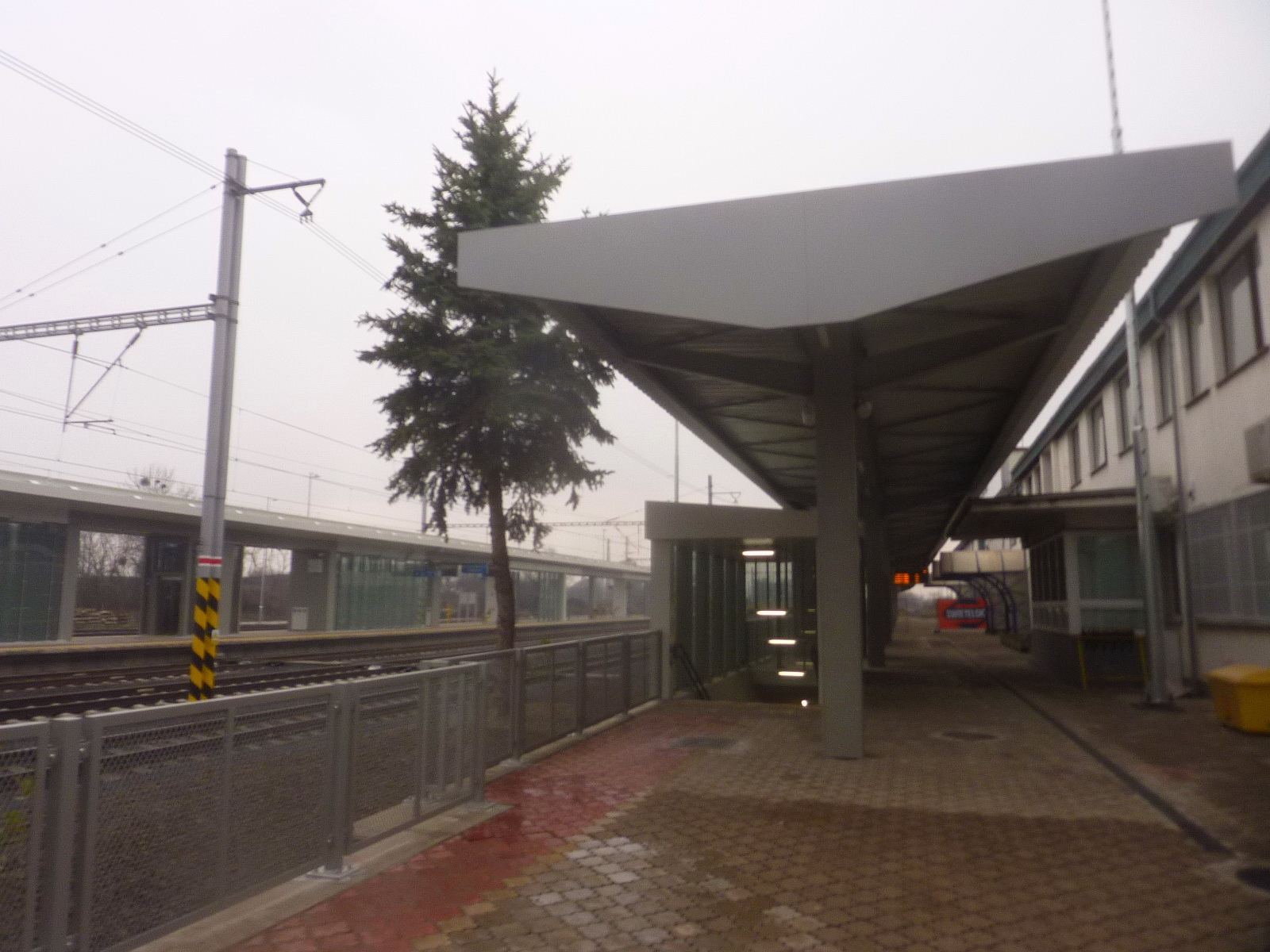 Quai de gare presque désert au petit matin, rails luisants et panneau lumineux allumé